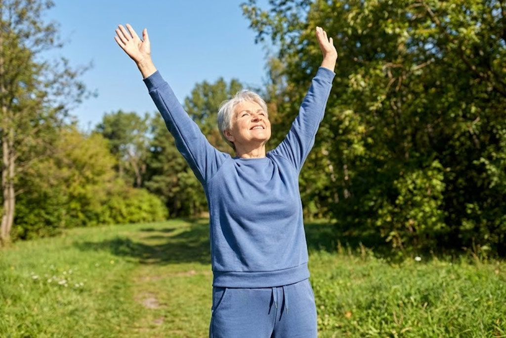 Senior femme souriante, debout dans un parc ensoleillé, les bras levés vers le ciel en train de s'étirer de tout son long pour lutter contre la raideur générale.