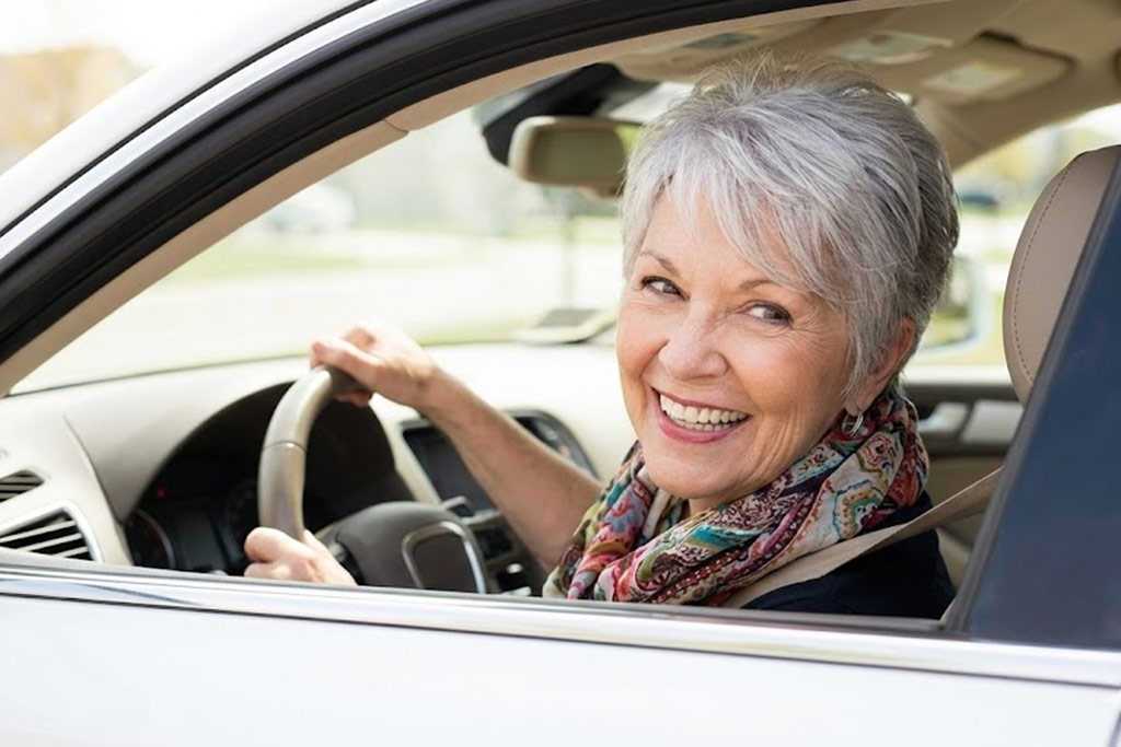 Senior femme souriante au volant de sa voiture, réussissant à tourner la tête pour regarder par-dessus son épaule sans douleur, dans un habitacle lumineux.