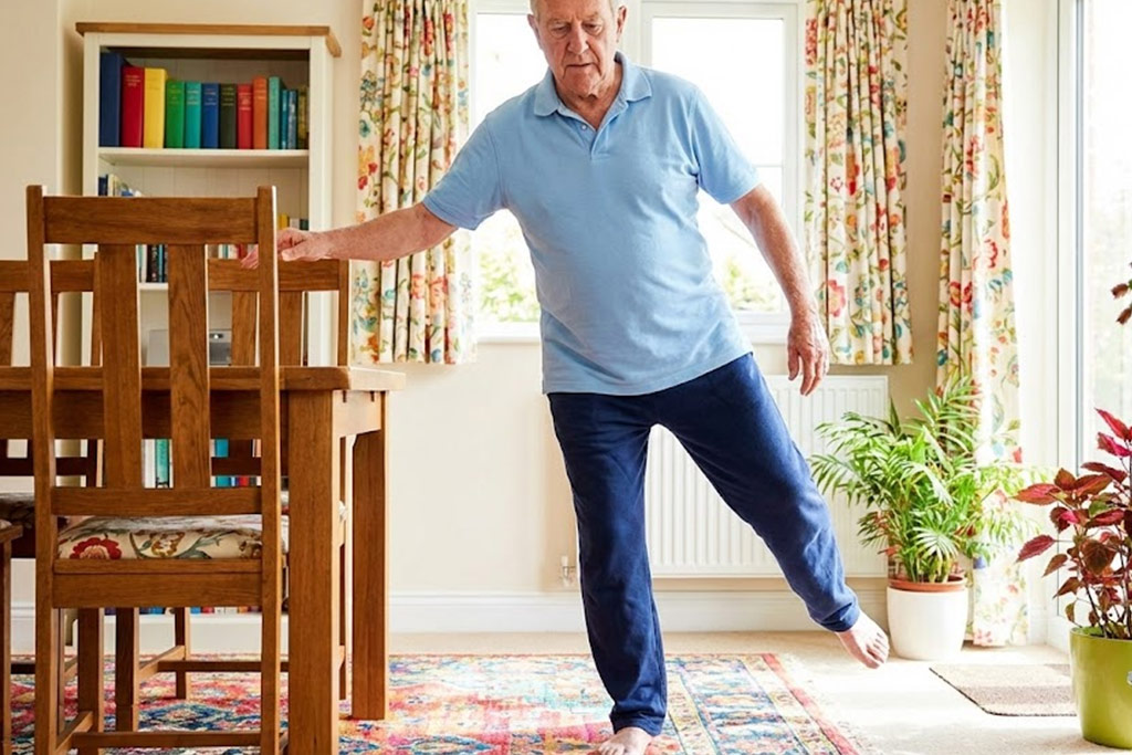 Homme senior concentré, se tenant légèrement au dossier d'une chaise dans un salon, effectuant l'exercice du flamant rose (debout sur une jambe) pour travailler son équilibre.
