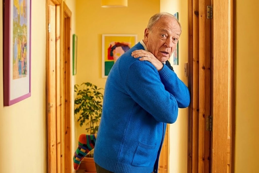 Senior homme avec une expression de surprise comique, se frottant l'épaule après s'être cogné dans le cadre d'une porte dans un couloir coloré. Cette image illustre avec humour les problèmes de coordination visuo-motrice ("syndrome du radar décalé") chez les seniors.